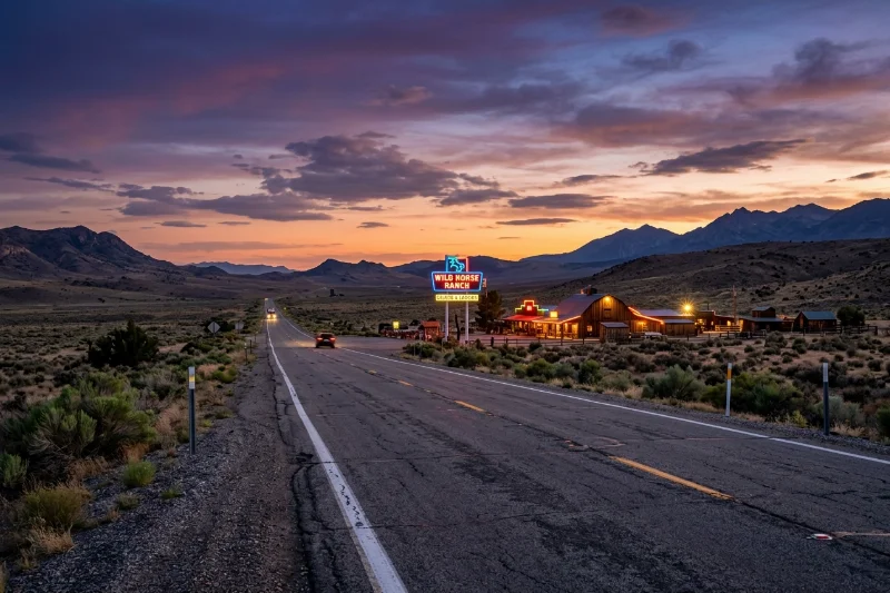 A desert road in Nevada leading toward a legal brothel ranch with neon signs under a sunset sky.